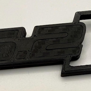 May include: Black 3D-printed keychain featuring the letters "SL" in a stylized font. The keychain includes a silver metal ring for attaching keys. The letters are thick and raised, with a textured surface.