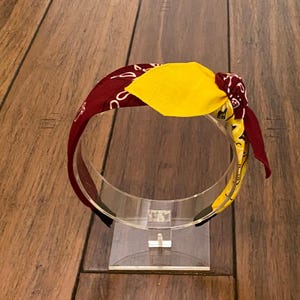 Op de afbeelding: Een rood en geel bandana-hoofdband met een knoop bovenop. De hoofdband staat op een transparante plastic standaard.