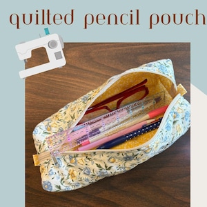 Puede incluir: Estuche acolchado con estampado floral, abierto para revelar lápices, una regla y gafas. También se ve el texto "easy instructions" y "by the bay bag co.". El estuche mide aproximadamente 23 cm de largo.