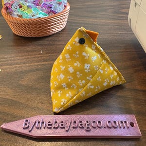 May include: A handmade chicken-shaped sewing accessory in yellow floral fabric with an orange beak and a black button eye. A basket of colourful sewing clips sits nearby. A pink sign reads "Bythebaybagco.com".