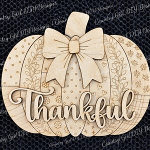 May include: A light-colored wooden pumpkin cutout featuring a layered bow at the top and the word Thankful in cursive script across the center. The pumpkin surface is divided into vertical sections with engraved patterns including dots, leaves, and hearts.