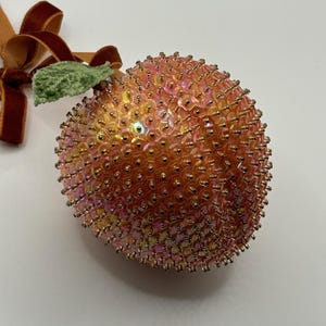 May include: A peach-coloured Christmas bauble covered in iridescent sequins with a green felt leaf and a brown velvet ribbon.