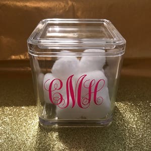Op de afbeelding: Een vierkante container van helder plastic met een deksel, gevuld met witte wattenbolletjes. De container heeft een roze monogram "CMH" aan de voorkant.