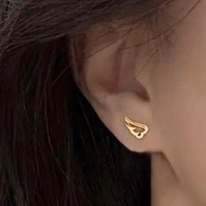 May include: A pair of gold wing-shaped earrings. The earrings are small and delicate, and they are worn in the wearer's earlobes.