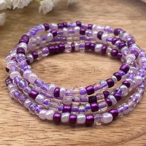 May include: Four beaded bracelets in shades of purple, lavender, and clear. The bracelets are made of small, round beads and are stacked on top of each other. The bracelets are on a wooden surface.