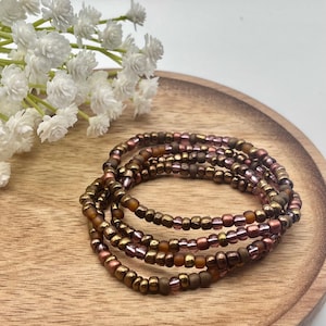 Könnte beinhalten: Eine Sammlung von Perlenarmbändern in Braun-, Gold- und Rosatönen. Die Armbänder liegen auf einem Holztablett, im Hintergrund weiße Blüten. Die Perlen haben einen metallischen Glanz.