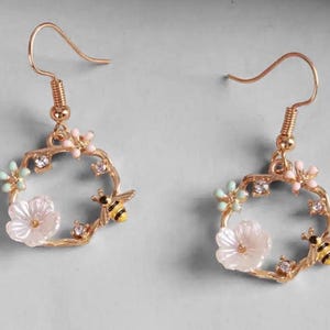 Pode incluir: Brincos de argola dourados com um design floral e de abelha. Os brincos apresentam um círculo de metal dourado com flores rosa, brancas e azuis e uma abelha com listras pretas e amarelas.