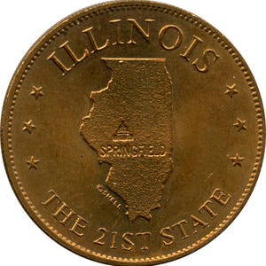 May include: A bronze-colored coin featuring the state of Illinois. The coin has the word "ILLINOIS" at the top, the state outline in the center, and "SPRINGFIELD" below it. The text "THE 21ST STATE" is at the bottom, with stars around the edge.