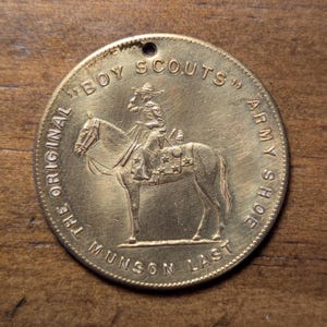May include: A gold-coloured metal token with the inscription "The Original Munson Last Army Shoe Boy Scouts" and a depiction of a person on horseback.