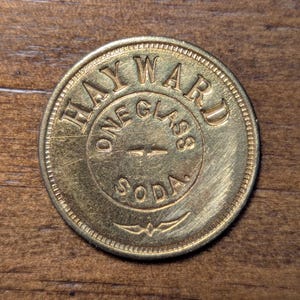 May include: A round, antique brass token with the embossed text "HAYWARD ONE GLASS SODA." The token has a detailed border and is set against a wooden surface. The text is in a classic, vintage font.