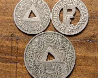 May include: Three vintage metal tokens. Two tokens are inscribed with "PACIFIC GAS AND ELECTRIC CO." and a triangle. The third token is inscribed with "PACIFIC ELECTRIC RAILWAY CO." and the letters "P" and "E".