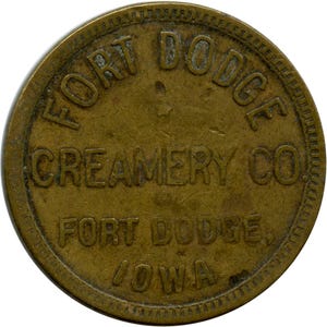 Puede incluir: Una ficha circular de latón con un borde elevado y una superficie texturizada. El texto FORT DODGE CREAMERY CO. está grabado en el centro, con FORT DODGE, IOWA debajo. El metal muestra signos de antigüedad y desgaste con una pátina oscura.