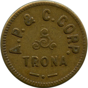 Op de afbeelding: Een ronde, bronskleurige munt met de tekst "A.P. & C. CORP. TRONA" en een decoratief wervelontwerp in het midden. De munt heeft een getextureerde rand en is waarschijnlijk een vintage token of munt.