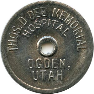 May include: A circular, metallic token with a dark, weathered finish and a central hole. The text embossed on the surface reads THOS. D DEE MEMORIAL HOSPITAL in an arc at the top, with OGDEN, UTAH printed in two lines below the center hole.