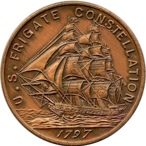 May include: A bronze coin depicting a sailing ship with detailed rigging and sails. The coin is inscribed with "U.S. FRIGATE CONSTELLATION" and the year "1797". The coin is circular and has a weathered, aged appearance.