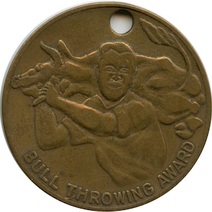 Könnte beinhalten: Runde, bronzefarbene Medaille mit einem Loch oben. Sie zeigt ein Relief einer Person, die einen Bullen wirft, mit den Worten "BULL THROWING AWARD" am unteren Rand. Das Design ist detailliert und zeigt die Figuren in Aktion.