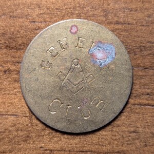 May include: A round, brass-colored token with the words "GENEL CLUB" and a Masonic symbol in the center. The token has a worn appearance with some discoloration and a small, pinkish spot near the top. The background is a wooden surface.