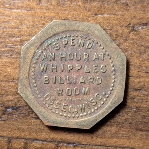 May include: A vintage, octagonal brass token with the text "SPEND AN HOUR AT WHIPPLES BILLIARD ROOM OSSEO, WIS." The token has a worn, aged appearance, suggesting it is an antique or collectible item.