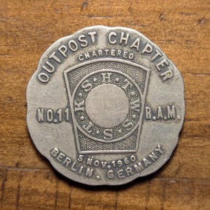 May include: A silver-coloured Outpost Chapter commemorative coin. The coin features the text "OUTPOST CHAPTER CHARTERED" around the edge, with "NO.11" and "R.A.M." on opposite sides. The centre has a shield design with the text "K.S.H.T.W.S.S." and "5 NOV.1960 BERLIN - GERMANY".