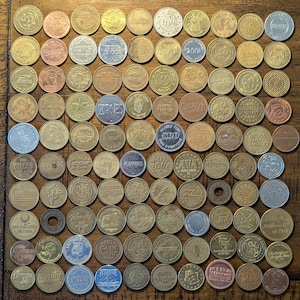 May include: A collection of assorted metal tokens, many with embossed text and designs. The tokens vary in color, including gold, silver, and bronze. Some tokens feature text like "ZONES", "FUN ZONE", and "FLIPPERS". The tokens are arranged on a wooden surface.