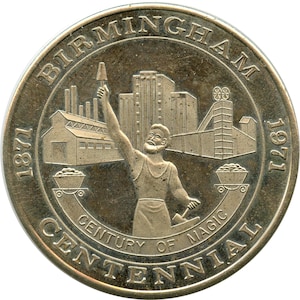 May include: A silver-colored commemorative coin with the text "BIRMINGHAM CENTENNIAL" and the years 1871 and 1971. The coin features a man holding a tool, industrial buildings, and the words "CENTURY OF MAGIC."