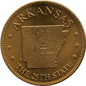 May include: A gold-colored coin featuring the outline of the state of Arkansas. The word "ARKANSAS" arches across the top, with "THE 25TH STATE" at the bottom. The city "LITTLE ROCK" is also visible.