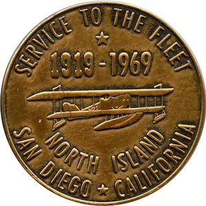 Puede incluir: Una moneda conmemorativa de color bronce con el texto "SERVICE TO THE FLEET" y "1919-1969" que rodea un diseño de hidroavión. La moneda también presenta "SAN DIEGO NORTH ISLAND CALIFORNIA".