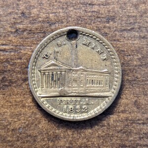 May include: A vintage brass coin featuring a building illustration and the text "U.S. MINT PHILA. 1882". The coin has a hole at the top and a rope-like border. It rests on a wooden surface.