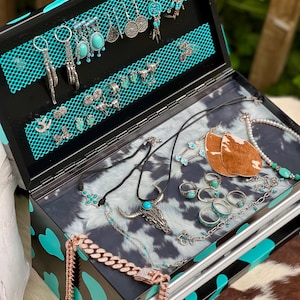 May include: Open black jewellery box with turquoise and silver earrings, necklaces, and rings. The box has a black and turquoise cow print design. The jewellery includes turquoise stones and silver accents, with a cowhide pattern inside.