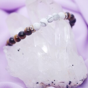 Puede incluir: Un brazalete de cuentas marrones y blancas con un dije de cristal. El brazalete está apoyado en un cristal blanco.