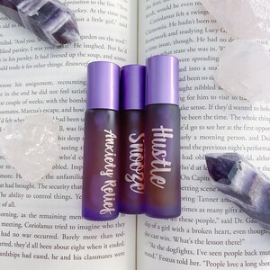Puede incluir: Tres botellas de vidrio transparente y morado con etiquetas que dicen "Hustle", "Snooze" y "Anxiety Relief". Las botellas están sobre una superficie blanca con un cristal morado.