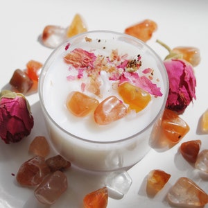 Puede incluir: Un portavelas de cristal transparente lleno de cera blanca, adornado con piedras preciosas naranjas, p&eacute;talos de rosa secos y un capullo de rosa rosa. La vela est&aacute; rodeada de m&aacute;s piedras preciosas naranjas sobre una superficie blanca.