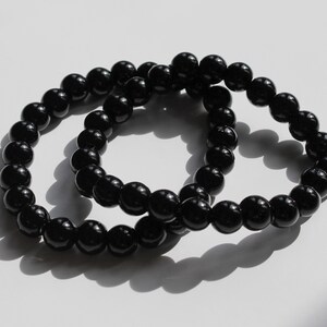 Puede incluir: Dos pulseras de cuentas negras, una ligeramente más grande que la otra, están entrelazadas y dispuestas sobre una superficie blanca.