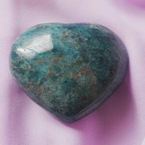 Puede incluir: Una piedra preciosa verde y gris en forma de corazón sobre un fondo de tela de color púrpura claro.
