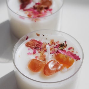 Puede incluir: Dos peque&ntilde;os portavelas transparentes llenos de cera blanca. Cada vela est&aacute; decorada con piedras preciosas naranjas y p&eacute;talos de rosa rosas secos. Las velas est&aacute;n sobre una superficie blanca.