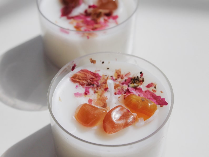 Puede incluir: Dos peque&ntilde;os portavelas transparentes llenos de cera blanca. Cada vela est&aacute; decorada con piedras preciosas naranjas y p&eacute;talos de rosa rosas secos. Las velas est&aacute;n sobre una superficie blanca.