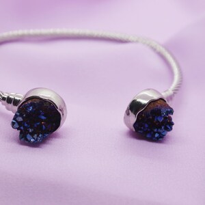 Puede incluir: Un brazalete de plata con dos acentos de cristal azul. Los cristales tienen forma de racimo y tienen un brillo metálico.