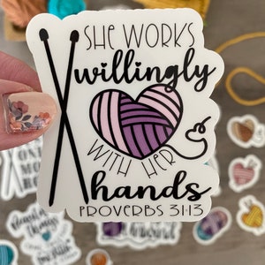 May include: A white sticker with the text "She works willingly with her hands" and "Proverbs 31:13". The sticker features a heart-shaped yarn ball and crossed knitting needles. Other stickers and yarn are in the background.
