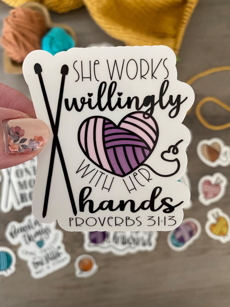 May include: A white sticker with the text "She works willingly with her hands" and "Proverbs 31:13". The sticker features a heart-shaped yarn ball and crossed knitting needles. Other stickers and yarn are in the background.
