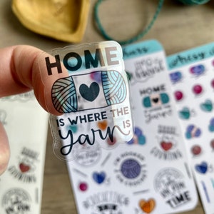 Peut inclure: Un autocollant transparent avec le texte "HOME IS WHERE THE YARN IS" et une pelote de laine color&eacute;e avec un c&oelig;ur au centre. L'autocollant est tenu par un doigt.