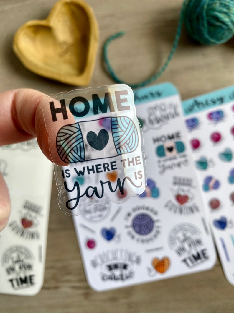 Peut inclure: Un autocollant transparent avec le texte "HOME IS WHERE THE YARN IS" et une pelote de laine color&eacute;e avec un c&oelig;ur au centre. L'autocollant est tenu par un doigt.