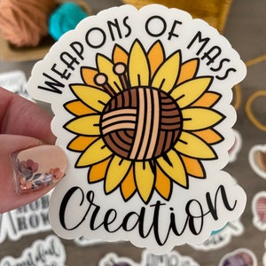 May include: A white sticker with the text "WEAPONS OF MASS CREATION" in black script. The sticker features a sunflower design with a yarn ball center and knitting needles. The petals are yellow and orange.