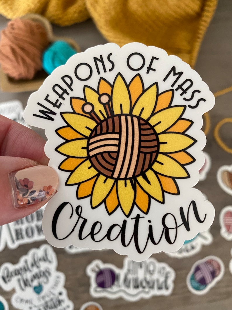 May include: A white sticker with the text "WEAPONS OF MASS CREATION" in black script. The sticker features a sunflower design with a yarn ball center and knitting needles. The petals are yellow and orange.