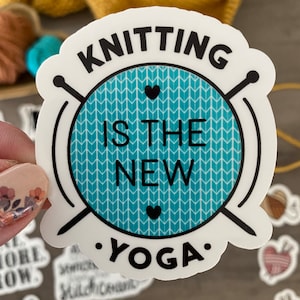 May include: A white sticker with the text "KNITTING IS THE NEW YOGA" in black. The text is arranged around a teal circle with a white chevron pattern and two black hearts. Knitting needles are on either side of the circle.