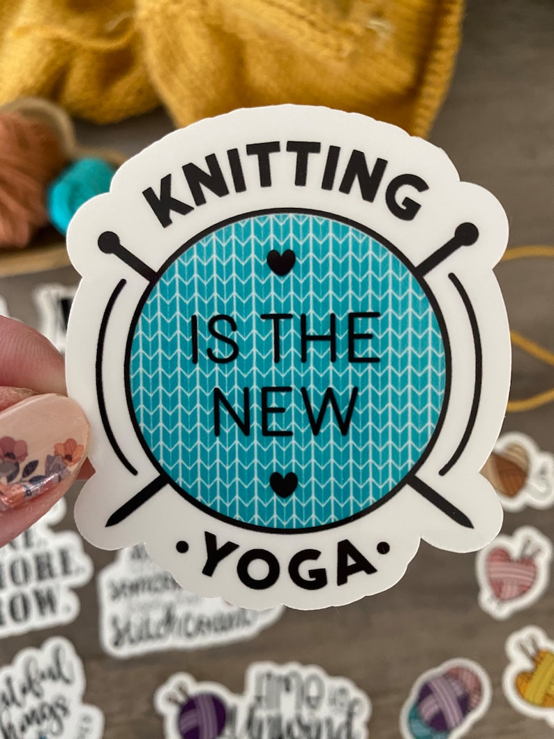 May include: A white sticker with the text "KNITTING IS THE NEW YOGA" in black. The text is arranged around a teal circle with a white chevron pattern and two black hearts. Knitting needles are on either side of the circle.