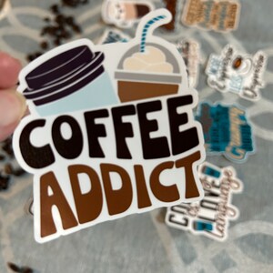 以下が含まれることがあります： 「COFFEE ADDICT」と茶色と黒の文字で書かれた白いステッカー。ステッカーには、蓋とストローが付いたコーヒーカップと、ホイップクリームとストローが付いた茶色のカップの漫画の画像が描かれています。