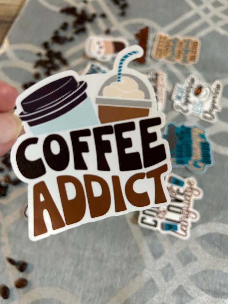 以下が含まれることがあります： 「COFFEE ADDICT」と茶色と黒の文字で書かれた白いステッカー。ステッカーには、蓋とストローが付いたコーヒーカップと、ホイップクリームとストローが付いた茶色のカップの漫画の画像が描かれています。