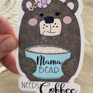 May include: A brown bear with a flower crown holding a blue mug with the text "Mama Bear Needs Coffee".
