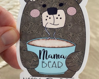 May include: A brown bear with a flower crown holding a blue mug with the text "Mama Bear Needs Coffee".
