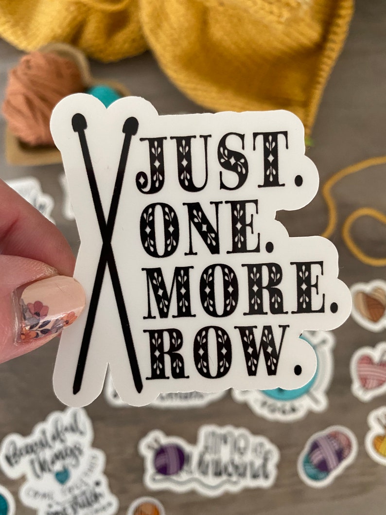 May include: White sticker with black text and crossed knitting needles. The text reads "JUST. ONE. MORE. ROW." Other stickers with knitting and crochet themes are in the background.